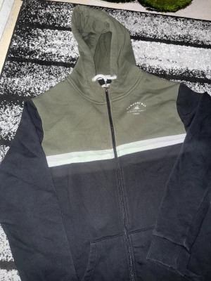 Grön och svart hoodie Newport Bay - Snygg hoodie från Newport Bay i storlek medium. Färgblockad design med olivgrön överdel, svart nederdel och vit rand över bröstet. Dragkedja framtill, fickor och justerbar huva med vita snören. Mjuk bomullskänsla och tryck på bröstet med seglingsmotiv.