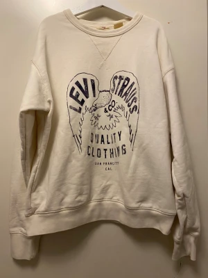 Vit sweatshirt från Levi's XS - Säljer en vit sweatshirt från Levi's i storlek XS. Tröjan har ett stort tryck med Levi Strauss & Co och en cool örn på framsidan. Rund hals och långa ärmar, tillverkad i mjuk bomull. Perfekt för dig som gillar streetwear och klassiska märken.