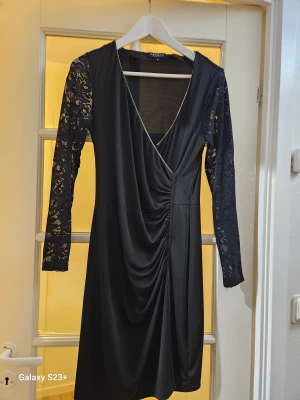 Svart omlottklänning från Morgan - Svart kort omlottklänning från Morgan med v-ringning och snygga spetsärmar. Klänningen har draperad front och lång ärm i transparent spets som ger en elegant touch. Perfekt för dig som vill sticka ut med stilren look.