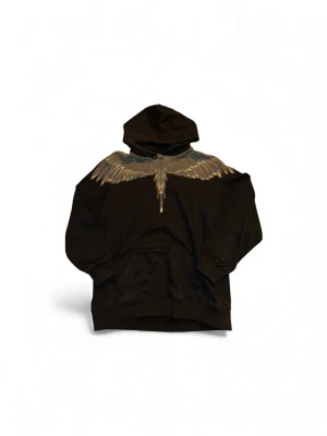 Svart hoodie Marcelo Burlon med vingar - Svart hoodie från Marcelo Burlon County of Milan med ikoniskt vingtryck i grått och guld över axlarna. Klassisk känguruficka framtill, huva med dragsko och ribbade muddar. Tillverkad i 100% bomull för en skön och avslappnad passform.
