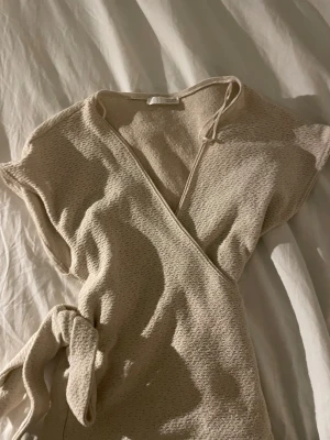 Beige omlottopp med knytband - Snygg beige omlottopp i stickad struktur med korta ärmar och knytband i midjan. Perfekt för lager på lager och ger en avslappnad men ändå stilren look. Materialet känns mjukt och luftigt, vilket gör den skön att bära under varmare dagar.
