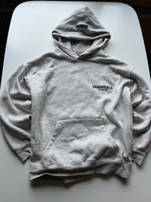Grå Essentials Fear of God hoodie - Snygg ljusgrå hoodie från Essentials Fear of God med svart logga på bröstet. Klassisk passform, stor huva och känguruficka framtill. Tillverkad i mjuk bomullsblandning som känns riktigt skön mot huden. Perfekt för en clean och avslappnad streetwear-look.