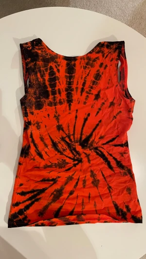 Orange och svart tie-dye linnetopp - Cool ärmlös linnetopp i röd och svart tie-dye med rundad hals och öppen. Toppens mönster är unikt och ger en edgy vibe, perfekt för att sticka ut. Tunn och mjuk, passar till jeans eller kjol för en trendig look😎