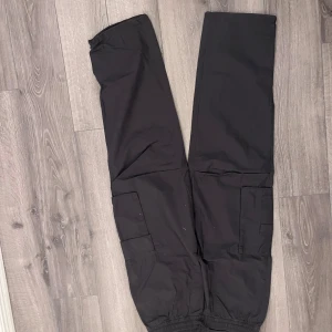 Svarta cargopants från H&M - Svarta cargopants från H&M med elastisk midja och stora fickor på sidorna. Byxorna har en loose passform och är tillverkade i ett syntetmaterial som känns lätt och smidigt. Perfekt för dig som gillar streetwear och funktionella detaljer.