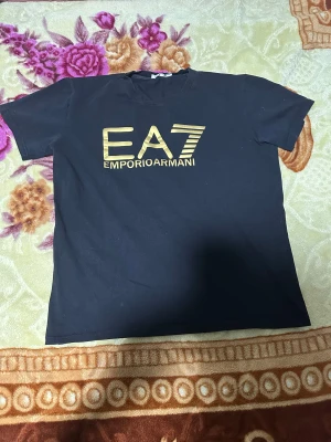 Svart EA7 Emporio Armani t-shirt - Svart t-shirt från EA7 Emporio Armani med guldig logga framtill. Klassisk passform med rund hals och korta ärmar. Tillverkad i mjuk bomull som känns skön mot huden. Perfekt för dig som gillar stilrena och exklusiva plagg.