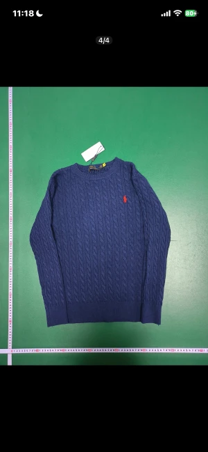 Blå stickad tröja Polo Ralph Lauren - Snygg marinblå kabelstickad tröja från Polo Ralph Lauren med rund hals och lång ärm. Tröjan har det klassiska röda Polo-logot broderat på bröstet och är tillverkad i mjuk bomull. Perfekt för dig som gillar stilrena och tidlösa plagg.