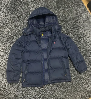 Mörkblå pufferjacka från Polo Ralph Lauren - Säljer en mörkblå pufferjacka från Polo Ralph Lauren i storlek XL barnstorlek. Jackan har huva, två sidofickor med dragkedja och klassisk röd Polo-logga på bröstet. Materialet är syntet och jackan är perfekt för kalla dagar. Stängs med dragkedja framtill.