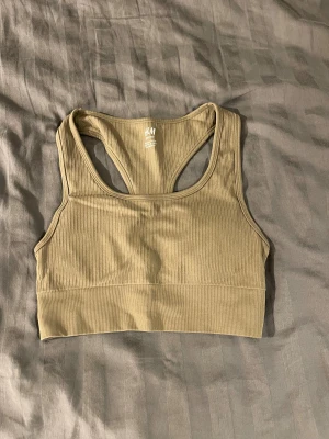 Ribbad beige sporttopp från H&M - Säljer en ribbad sporttopp från H&M i beige färg. Toppen har bred resår nedtill, brottarrygg och är ärmlös. Perfekt för träning eller chill dagar. Materialet är stretchigt och bekvämt, och modellen sitter tight mot kroppen.