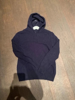 Mörkblå cashmere hoodie från Sayless - Snygg mörkblå hoodie från Sayless i mjuk och lyxig cashmere. Tröjan har huva med snörning och långa ärmar, perfekt för dig som gillar stilrena och bekväma plagg. Ribbstickade muddar vid ärmslut och nederkant ger en schysst passform. Den är XXS men passar XS