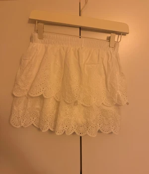 Vit volangkjol med broderade detaljer - Supersöt vit kortkjol med två lager volanger och broderade spetsdetaljer. Kjolen har elastisk midja och ett romantiskt, luftigt uttryck med scallop-kant. Perfekt för dig som gillar en feminin och fräsch stil.