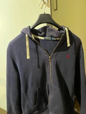 Mörkblå hoodie från Polo Ralph Lauren - Snygg mörkblå hoodie från Polo Ralph Lauren med dragkedja och huva. Tröjan har vita snören, röd broderad logga på bröstet och praktiska fickor framtill. Perfekt för chill dagar och enkel att matcha med jeans eller joggers.