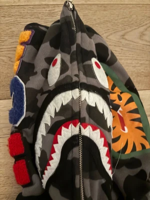 BAPE Shark Camo Zip Hoodie - Säljer en BAPE Shark Zip Hoodie med grå och svart camomönster, ikonisk hajdesign på huvan med broderade tänder och ögon, samt tigertryck på sidan. Färgglada bokstäver på huvan och klassisk BAPE-logga på mudden. Tillverkad i mjuk bomull med dragkedja hela vägen. Med en liten fläck där bak därför så lågt pris strlk s-M