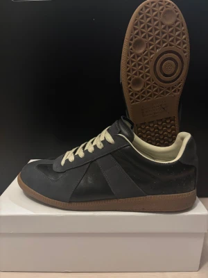 Maison Margiela mörkblå sneakers - Maison Margiela sneakers i mörkblå skinn och mocka med beige snörning och gummisula. Klassisk rund tå och platt sula, med ikonisk Margiela-etikett på tungan. Snygg kontrast mellan mörka paneler och ljus insida, perfekt för dig som gillar stilrena och exklusiva sneakers.