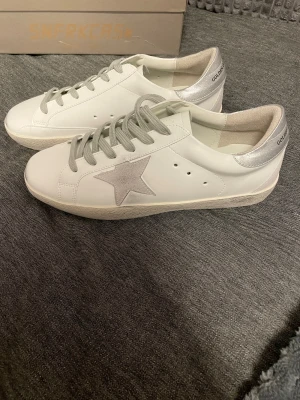 Golden Goose Superstar vita sneakers - Golden Goose Superstar sneakers i vitt skinn med silverfärgade detaljer och grå snörning. Sidan har en ikonisk stjärna i ljusgrå mocka och texten GGDB. Platt sula och rund tå. Insidan är märkt med 'SUPERSTAR'. Snygg och clean design för dig som gillar stilrena sneakers.