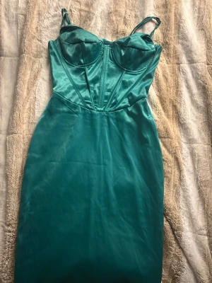 Grön satin klänning med bygel - Snygg grön klänning i satin med figurnära passform och justerbara smala axelband. Klänningen har bygel och markerade sömmar över bysten för en cool bustier-look. Perfekt för dig som vill sticka ut med en elegant och modern stil.