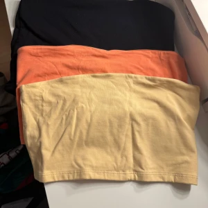 3-pack tubtoppar i olika färger - Tre stilrena bandeau tubtoppar i svart, orange och ljusgul. Perfekta att matcha med jeans eller kjol för en enkel och trendig look. Tillverkade i mjuk bomull med stretch för skön passform. Säljes som paket.