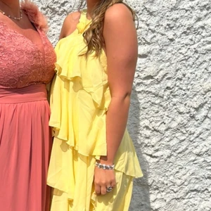 Gul långklänning med volanger - Supersöt gul långklänning med flera lager volanger och en snygg omlott vid halsen som knyts med en blomdetalj. Med öppen rygg. Perfekt till bal eller bröllop. Använd 1gång på ett bröllop 