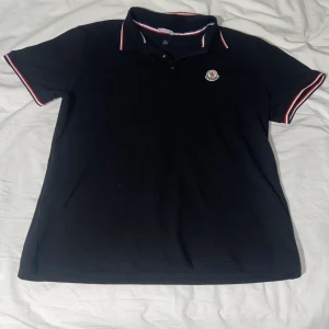 Svart pikétröja från Moncler - Snygg svart pikétröja från Moncler med klassisk krage och knäppning framtill. Tröjan har Moncler-logga på bröstet och röda, vita och blå detaljer på krage och ärmslut. Perfekt för en stilren och sportig look.