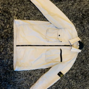 Vit overshirt från Stone Island - Snygg vit overshirt från Stone Island. Storlek M men passar även S. Relativt bra skick delvist några små fläckar men som försvinner ur tvätten! Vid minsta fundering är det bara o höra av sig!👊