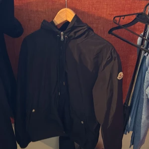 Svart vindjacka från Moncler - Svart vindjacka från Moncler med huva som har röd, vit och blå rand. Jackan har dragkedja framtill, två fickor med tryckknappar och Moncler-logga på ärmen. Perfekt för dig som vill ha en stilren och sportig look. Skick 9/10 pris kan diskuteras 