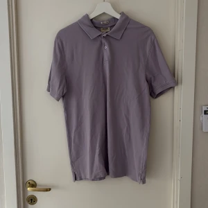 Ljuslila pikétröja från East West - Säljer en stilren ljuslila pikétröja från East West i ekologisk bomull. Tröjan har klassisk krage, kort ärm och knäppning framtill. Perfekt för en chill och fräsch look under vår och sommar.