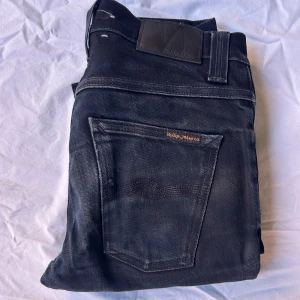 Svarta Nudie Jeans Grim Tim 28/30 - Svarta jeans från Nudie Jeans modell Grim Tim, tillverkade i 99% ekologisk bomull. Klassisk femficksdesign, knappgylf och diskreta kontrastsömmar. Snyggt broderad bakficka och logga på baklappen. Perfekta för dig som gillar stilrena svarta jeans.