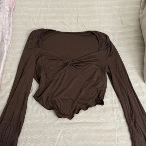 Brun croppad långärmad topp - Snygg brun långärmad topp med twistad detalj framtill och croppad passform. Toppen har en lätt vågig nederkant och är tillverkad i ett mjukt, stretchigt material. 