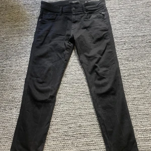 Svarta Replay jeans Anbass 32/30 - Svarta jeans från Replay, modell Anbass, i klassisk femficksdesign. Byxorna har smal passform och raka ben, tillverkade i mjukt bomullsmaterial med stretch för extra komfort. Perfekta för dig som gillar stilrena och enkla svarta jeans.