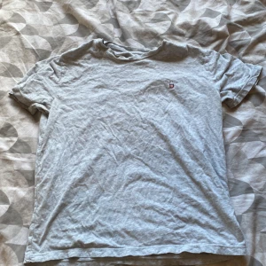 Ljusgrå t-shirt från GANT - Snygg ljusgrå t-shirt från GANT med klassisk logga och sköld på bröstet. T-shirten är i mjuk bomull och har en enkel, stilren design med korta ärmar och rund hals. Perfekt för en chill och avslappnad look.