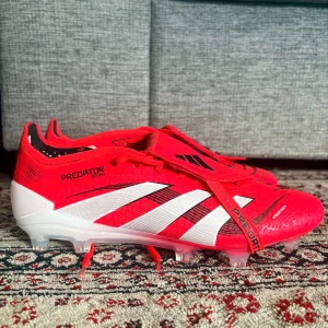 Adidas Predator  - Säljer ett par Adidas Predator fotbollsskor i rött och vitt med svarta detaljer. Skorna har snörning, klassiska tre ränder på sidan och mönstrad ovandel. Utrustade med dobbar för gräsplan och Predator-tryck under sulan. Perfekta för dig som vill sticka ut på planen.