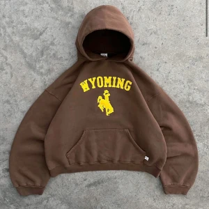 Brun Wyoming hoodie med tryck - Säljer en brun hoodie med gul text och motiv från Wyoming. Tröjan har huva, magficka och en oversized passform. Perfekt för dig som gillar college-stil och vill ha något bekvämt och snyggt.