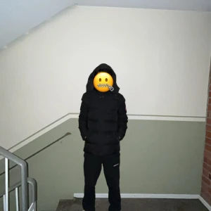 Svart dunjacka från Canada Goose - Säljer en svart dunjacka från Canada Goose med huva och klassisk logga på ärmen. Jackan är vadderad och har dragkedja samt knappar framtill. Perfekt för kalla dagar och har en clean, stilren look. Fint på folk som har S och perfekt på dom som har M.