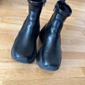 Buffalo svarta chunky boots i  (skinn oäkta)  - Svarta boots från Buffalo med chunky sula och dragkedja på sidan. Tillverkade i skinn med grovt mönstrad plattformssula för extra attityd. Klassisk rund tå och högt skaft, perfekta för dig som vill sticka ut med en edgy look.
