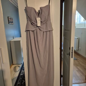 Ljuslila långklänning från TFNC - Elegant ljuslila långklänning från TFNC med axelbandslös design och draperad överdel. Klänningen har en vacker fallande kjol och är perfekt för speciella tillfällen. Tillverkad i ett lätt och luftigt material som ger en lyxig känsla.
