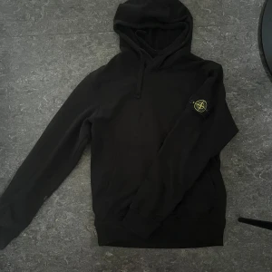 Svart hoodie från Stone Island - Svart hoodie från Stone Island med klassisk patch på vänster ärm. Tröjan har huva med snörning, känguruficka och långa ärmar. Tillverkad i mjuk bomull, perfekt för en chill och stilren look.