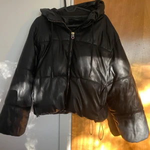 Svart pufferjacka från Bershka - Svart pufferjacka från Bershka med croppad passform och huva. Jackan har quiltad design, dragkedja framtill och snörning vid nederkanten. Materialet är syntet med skinnliknande yta som ger en cool vibe. Perfekt för dig som gillar streetstyle och vill ha något som sticker ut.