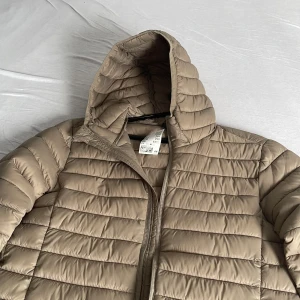 Beige pufferjacka med huva från H&M - Snygg beige pufferjacka från H&M med huva och horisontella sömmar. Jackan har dragkedja framtill och är tillverkad i ett lätt, syntetiskt material som håller dig varm. Perfekt för kyliga dagar och enkel att matcha med olika outfits.