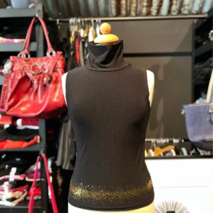 Svart polotopp med guldglitter  - Svart ärmlös polotopp från Koola Anna i stretchigt polyestermaterial. Toppen har hög krage och är dekorerad med guldglittrande detaljer längst ner. Snygg och figurnära passform som ger en cool look. Det är en liten M