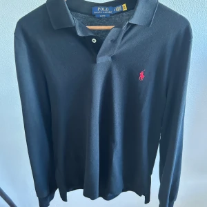 Svart långärmad pikétröja Polo Ralph Lauren - Svart långärmad pikétröja från Polo Ralph Lauren i slim fit-modell. Klassisk krage, knäppning framtill och ikonisk röd broderad logga på bröstet. Tillverkad i mjuk bomull som känns skön mot huden. Perfekt för en clean och stilren look.