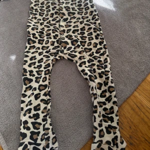 Leopardmönstrad byxdress i bomull - Byxdress med hel leopardmönster i beige, svart och brunt. Mjukt bomullsmaterial, långa ben och tryckknappar längs insidan för enkel påklädning. Perfekt för dig som gillar djurmönster och vill ha något bekvämt och snyggt.