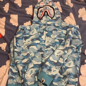 BAPE blå camo shark hoodie - Säljer en ikonisk BAPE shark hoodie i blå camo med dragkedja hela vägen upp till huvan. Tröjan har broderade detaljer, färgglada bokstäver och tiger-motiv på huvan. Perfekt statement-plagg för streetwear-stilen.
