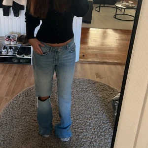 Ljusblå bootcut jeans med hål - Säljer ett par ljusblå jeans med bootcut-modell och låg midja. Jeansen har ett stort hål på ena knät för en extra edgy look. Klassisk femficksdesign och tillverkade i jeansmaterial. Perfekta för dig som gillar avslappnad och trendig stil.