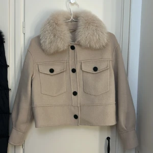 Beige jacka med pälsig krage - Säljer en beige jacka med stor fluffig pälsig krage och svarta knappar framtill. Jackan har två stora bröstfickor med lock och en croppad passform. Perfekt för dig som vill sticka ut med en snygg och trendig look.