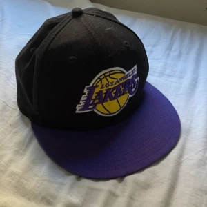 Los Angeles Lakers keps från New Era - Snygg Los Angeles Lakers keps från New Era 9FIFTY med svart krona och lila skärm. Lakers-logga broderad framtill i gult, lila och vitt. Kepsen är tillverkad i 100% bomull och har klassisk snapback-modell. Perfekt för basketfans som vill sticka ut.