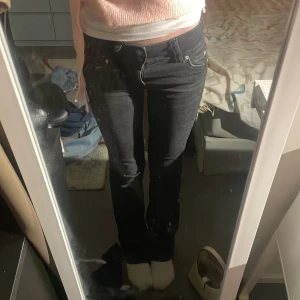 Svarta bootcut jeans från Bershka - Svarta jeans från Bershka i storlek 34 med bootcut passform. Jeansen har snygga bakfickor med lock och knappdetalj, klassisk femficksdesign och låg midja. Perfekta för dig som gillar en stilren look med lite edge.