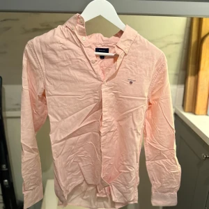 Ljusrosa skjorta från Gant - Säljer en ljusrosa skjorta från Gant med broderad logga. Skjortan har klassisk krage med knappar, lång ärm och är tillverkad i mjuk bomull. Perfekt för en clean och stilren look. 