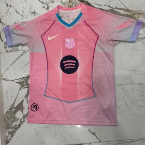 Rosa FC Barcelona x Nike fotbollströja - Snygg FC Barcelona fotbollströja från Nike i rosa och lila toner med vita detaljer. Tröjan har korta ärmar, V-ringad krage med blå kant och tryckta loggor från både Nike och Spotify. Tillverkad i lätt och ventilerande Dri-FIT-material. Storlek L men passar en lite större M