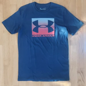 Blå Under Armour t-shirt  - Snygg marinblå t-shirt från Under Armour med stor logga och text på bröstet i blå och rött. Storlek small/medium. Tillverkad i mjuk bomullsmix, med en avslappnad stil. Perfekt för dig som gillar sportiga vibes.