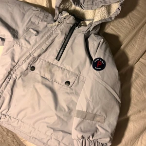 Ljusgrå vindjacka från Polarn O. Pyret - Säljer en ljusgrå vindjacka från Polarn O. Pyret med mysigt vitt fleecefoder i huvan. Jackan har en sned dragkedja, stor ficka framtill med tryckknappar och ett snyggt märke på ärmen. Perfekt för kyliga dagar och enkel att dra över huvudet. Skulle personligen säga att det är en vinterjacka! 