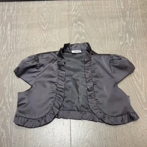 Grå bolero från Orsay i satin - Kort grå bolero från Orsay i glansigt satinmaterial. Boleron har volangdetaljer längs kanterna och vid kragen, samt korta ärmar. 